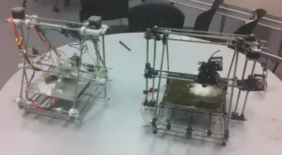 Image of two Prusa Mendel 3D printers on a desk. These printers have a trapezoid frame made from threaded rods and held in place with 3d printed pieces.
