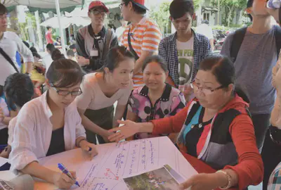 Residents, students and researchers gathered together. A few seated at a table. A resident holding images of the original space. A student drawing out a map and noting down comments.