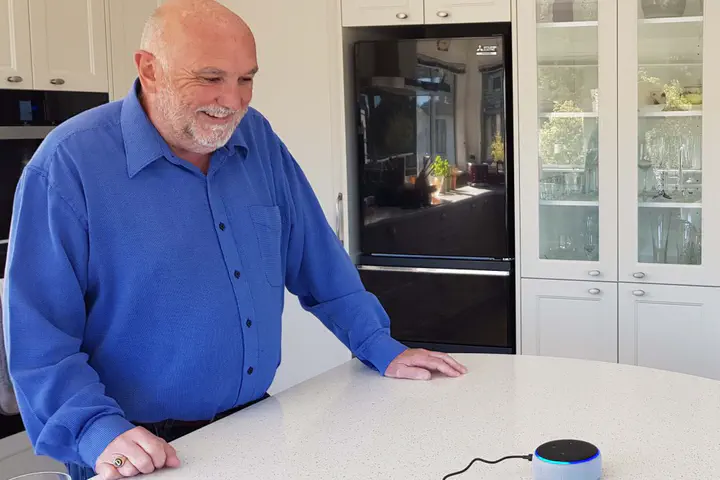 Older adult smiling and interacting with KinVoice from an Amazon Echo smart speaker device on the kitchen table